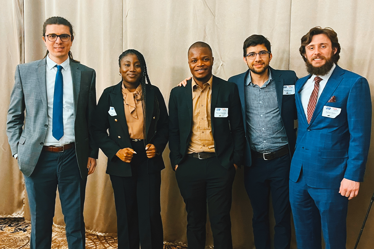 Five graduate students in business attire in front of golden curtain backdrop.