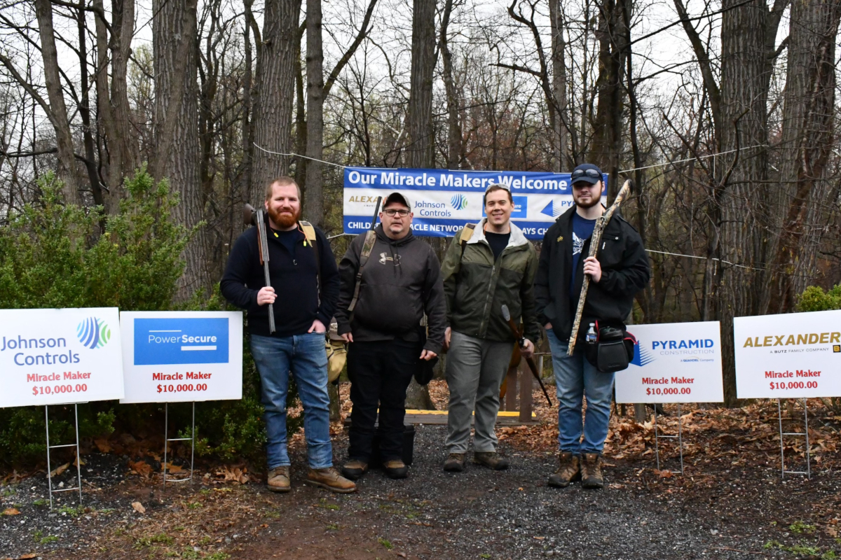 Four men pose for a photo at the edge of a wooded area. Three of them hold guns pointed at the ground.