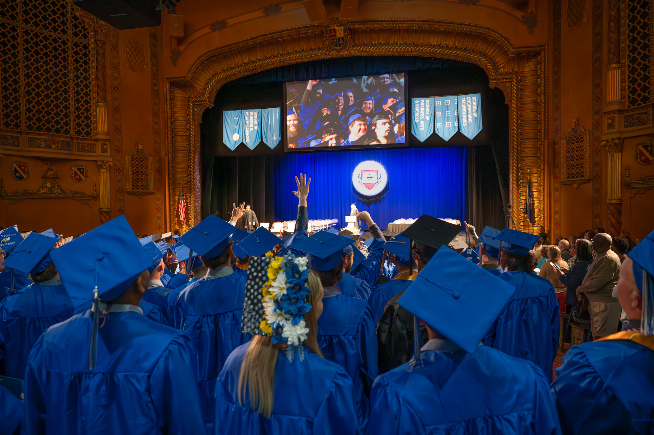 A view of the stage decorated for commencement over a sea of students wearing blue graduation mortarboard caps