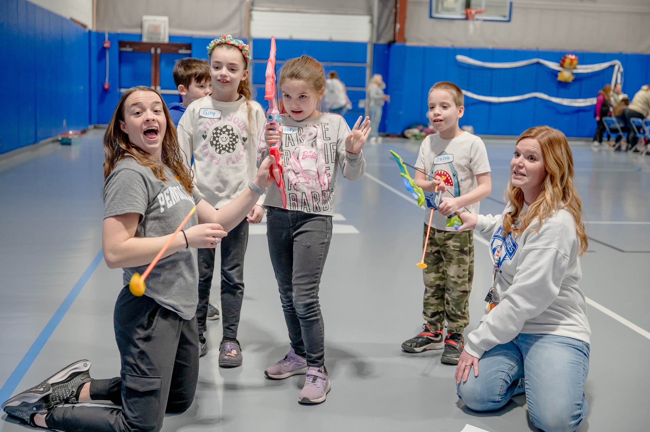 A couple of students help some children with a bow and arrow in a gymnasium