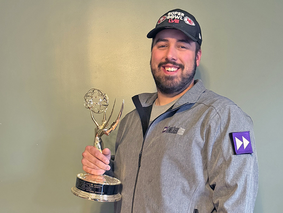 A man in a black baseball cap and gray jacket holding an Emmy statuette.