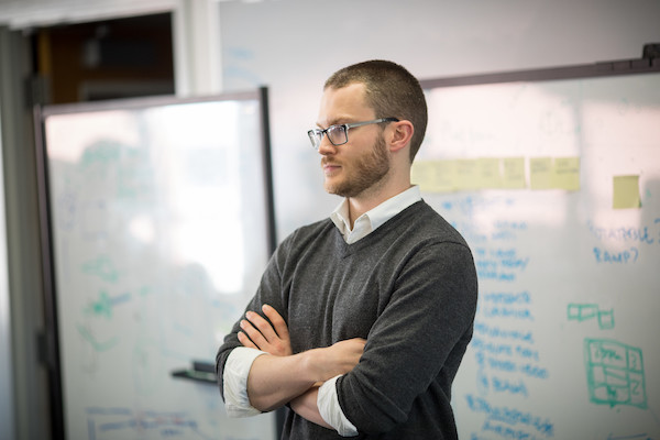 A professor stands with his arms crossed in front of whiteboards. 