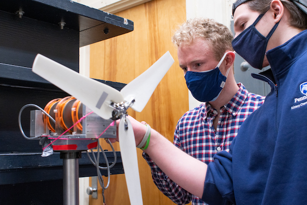 Two young men wearing face masks assemble a small wind turbine, which includes a motor, several cords and a white-winged propeller. 