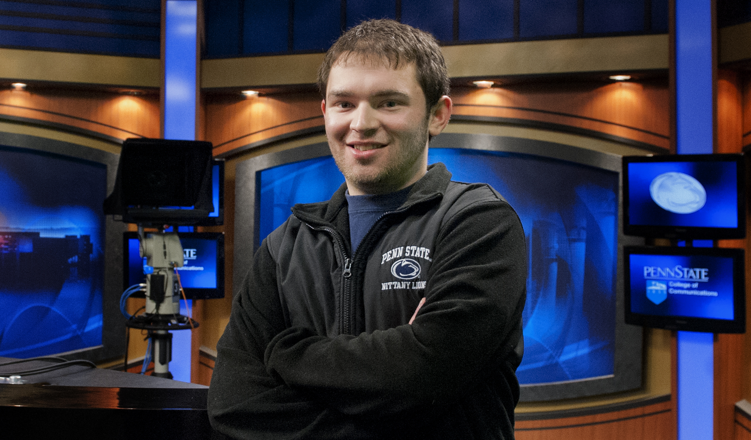 male college student standing with arms crossed in front of television camera recording equipment, studio set and TV monitors 