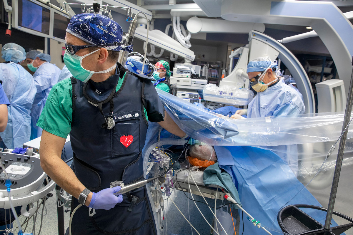 In the foreground, a surgeon watches a monitor while a second surgeon, background, prepares to insert a cardiovascular device on a patient