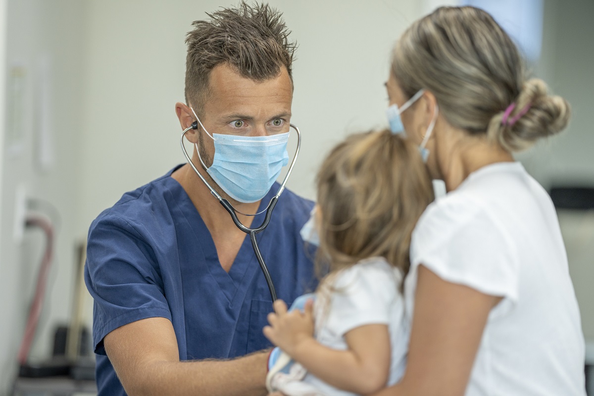 A nurse checks on a young patient's heartbeat. The little girl sits on her mother's lap while the nurse does the checkup.