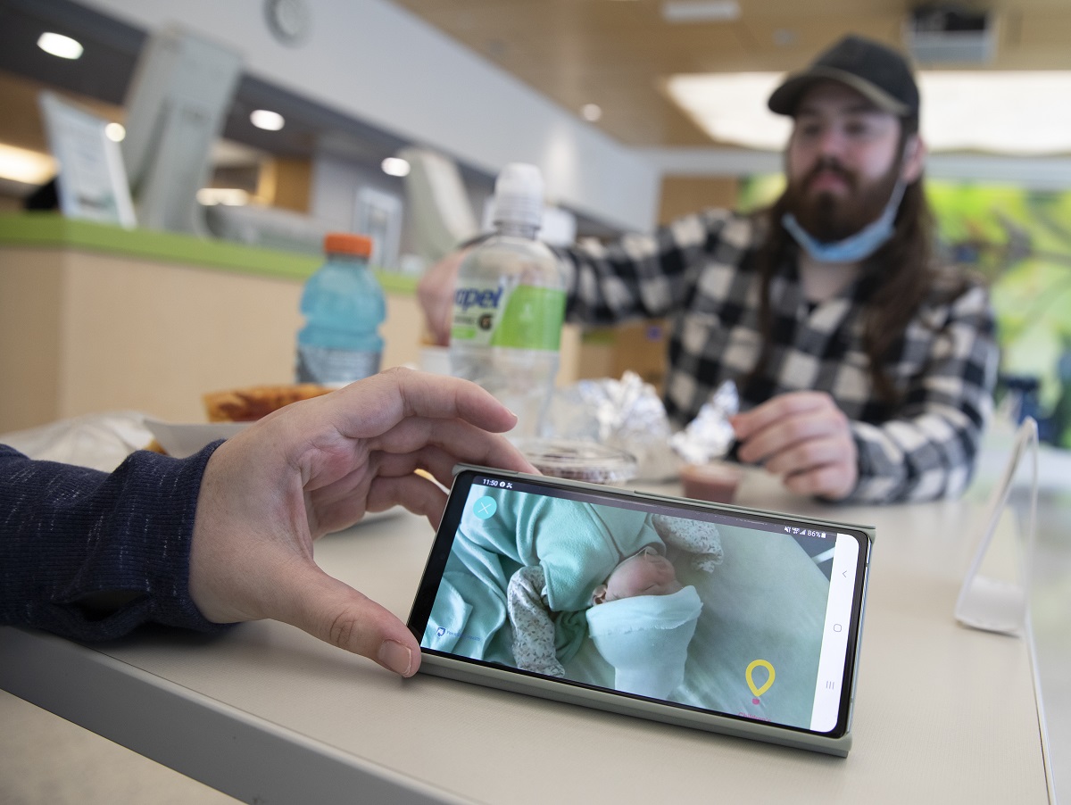 A hand holds a smart phone upon which is the image of an infant in a bed. Across a lunch table littered with bottles and food, a man with a beard and a hat looks on out of focus.