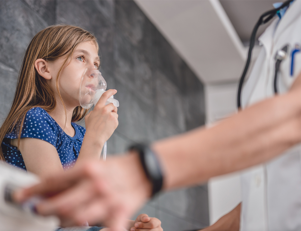 A young girl, seated, holds an oxygen mask up to her mouth and nose. A medical professional, in a white coat and wearing a stethoscope, is to the right, slightly out of focus.