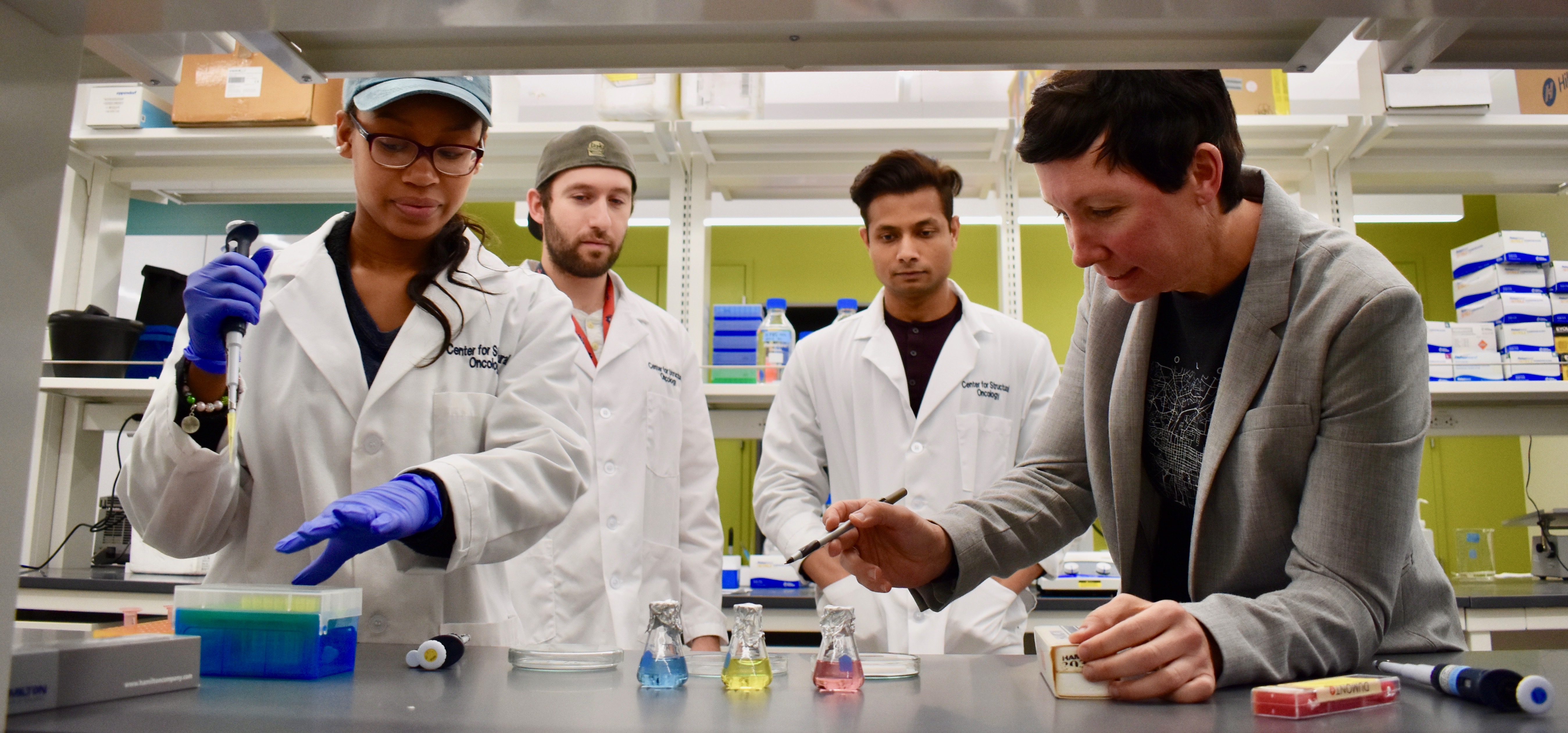 Two women and two men stand at a lab bench, looking at flasks containing different colored liquids.