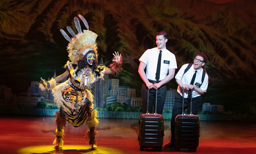 A person dressed in a costume featuring straw, beads and feathers gestures at two men wearing matching button-down shirts and slacks and toting luggage.