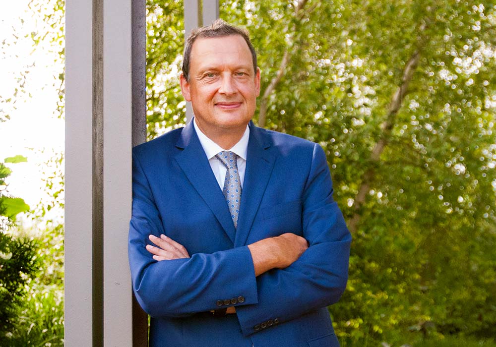 Michael Büsges in a blue suit with his arms crossed. Green foliage is in the background.