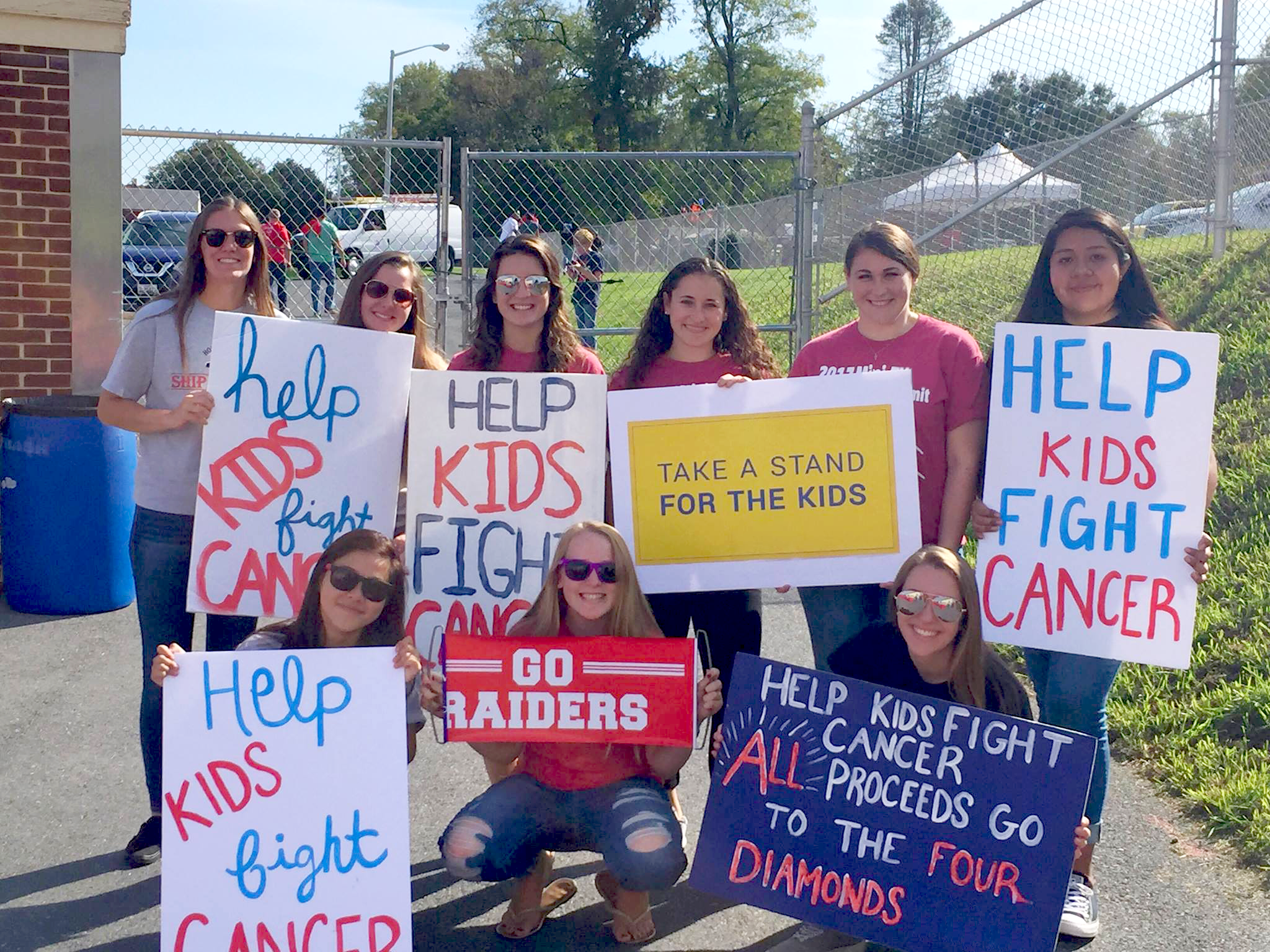 Nine young women pose outdoors, holding signs with slogans such as “Help Kids Fight Cancer” and “Take A Stand For The Kids.”