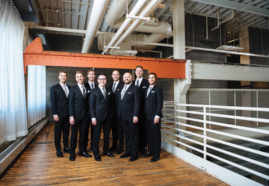 A group of men wearing the same suits stand in a cluster in a chic industrial surrounding.