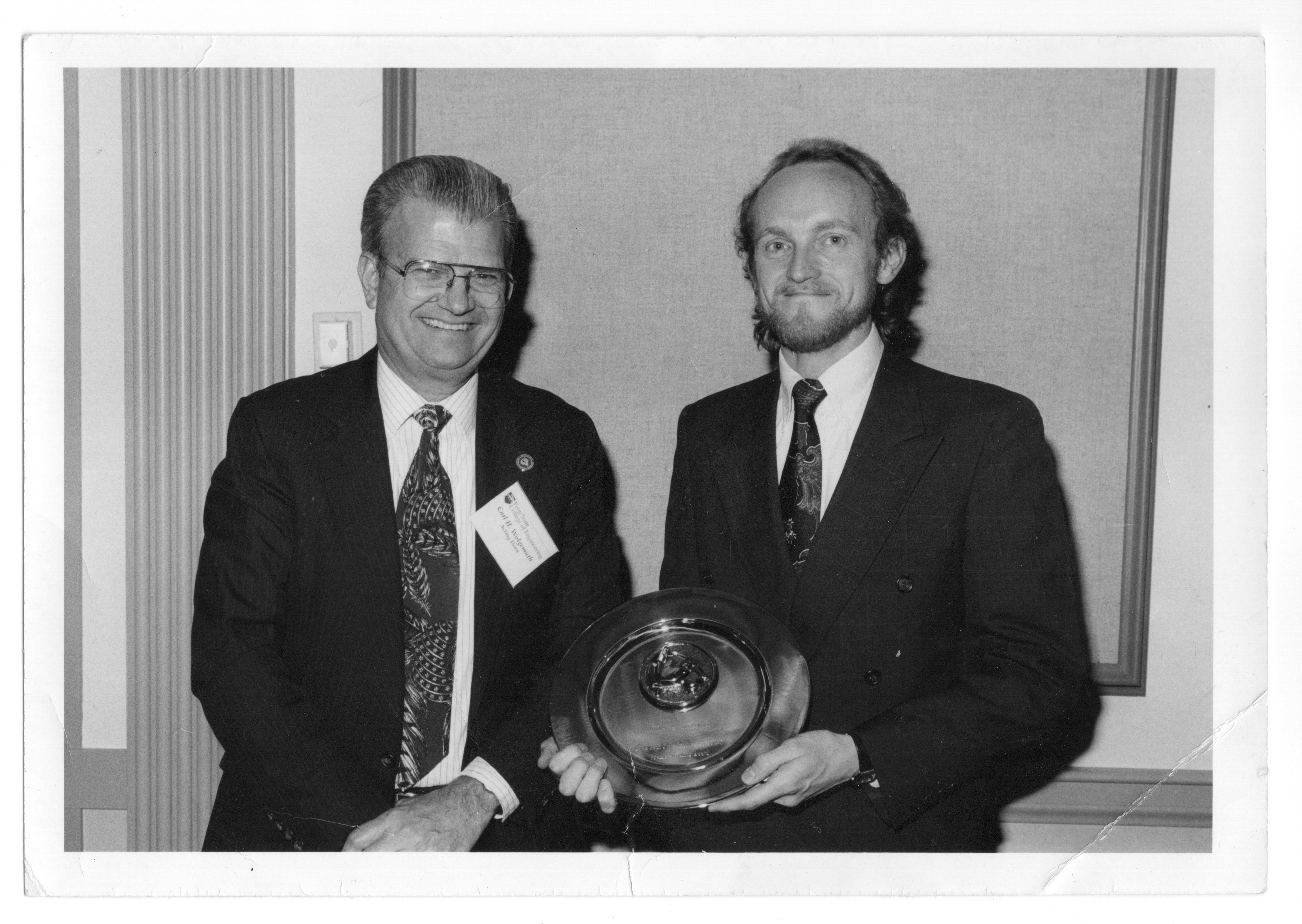 black and white photo of two people in suit and ties posing with an award