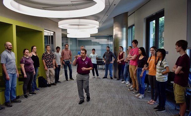 Man in red shirt standing in center of circle of students and gesturing with hands.