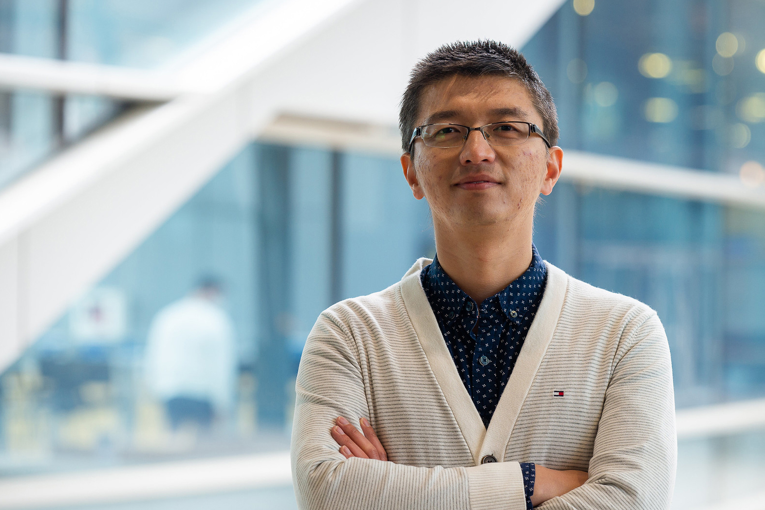 A man wearing glasses and a white cardigan poses for a photo in front of a window.