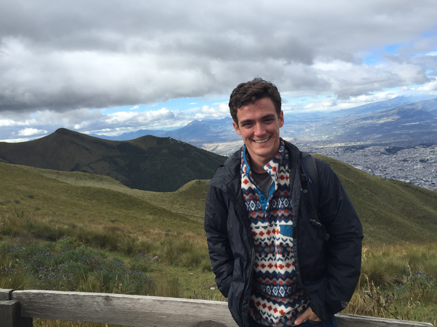 Matthew Crager stands in front of a small fence, smiling for the camera. Green mountains are seen in the distance. 