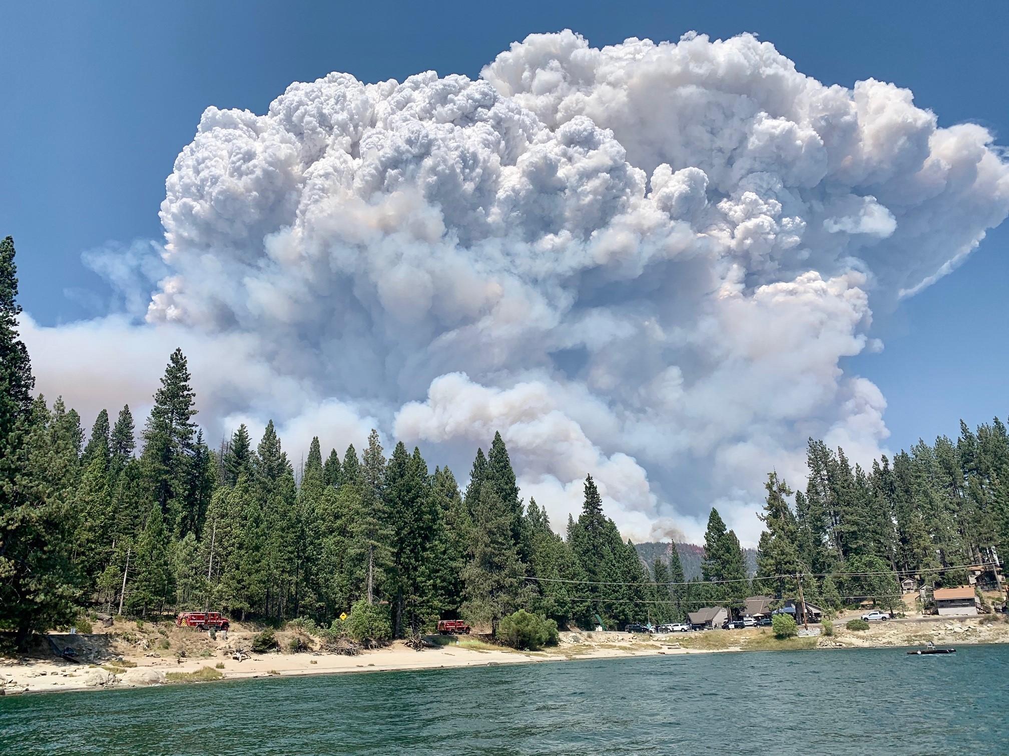 A lake and pine forest with a large cloud of smoke from a wildfire filling the sky