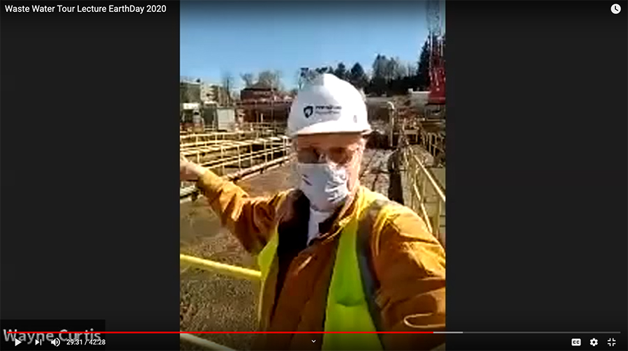 A man wearing a mask, hard hat, and safety glasses gestures to outdoor water pools in a screenshot of a YouTube video.