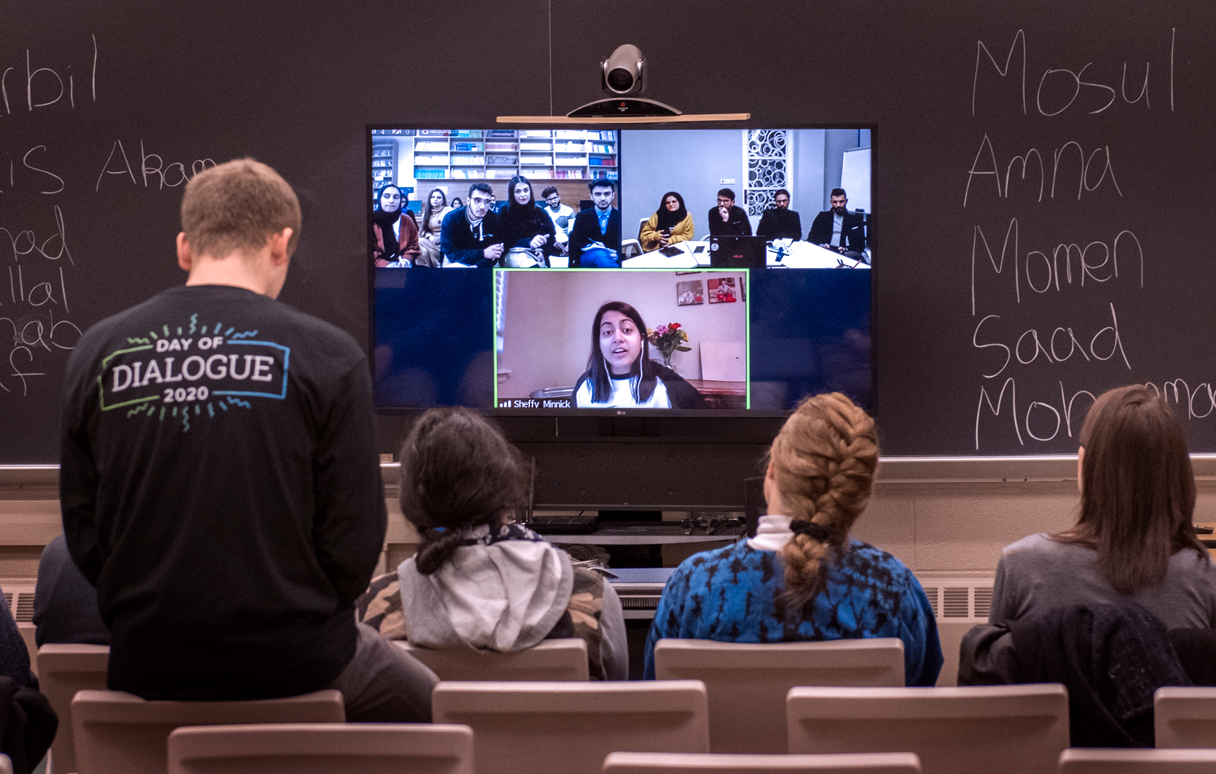 A group of people interacts with a woman who is speaking on a monitor