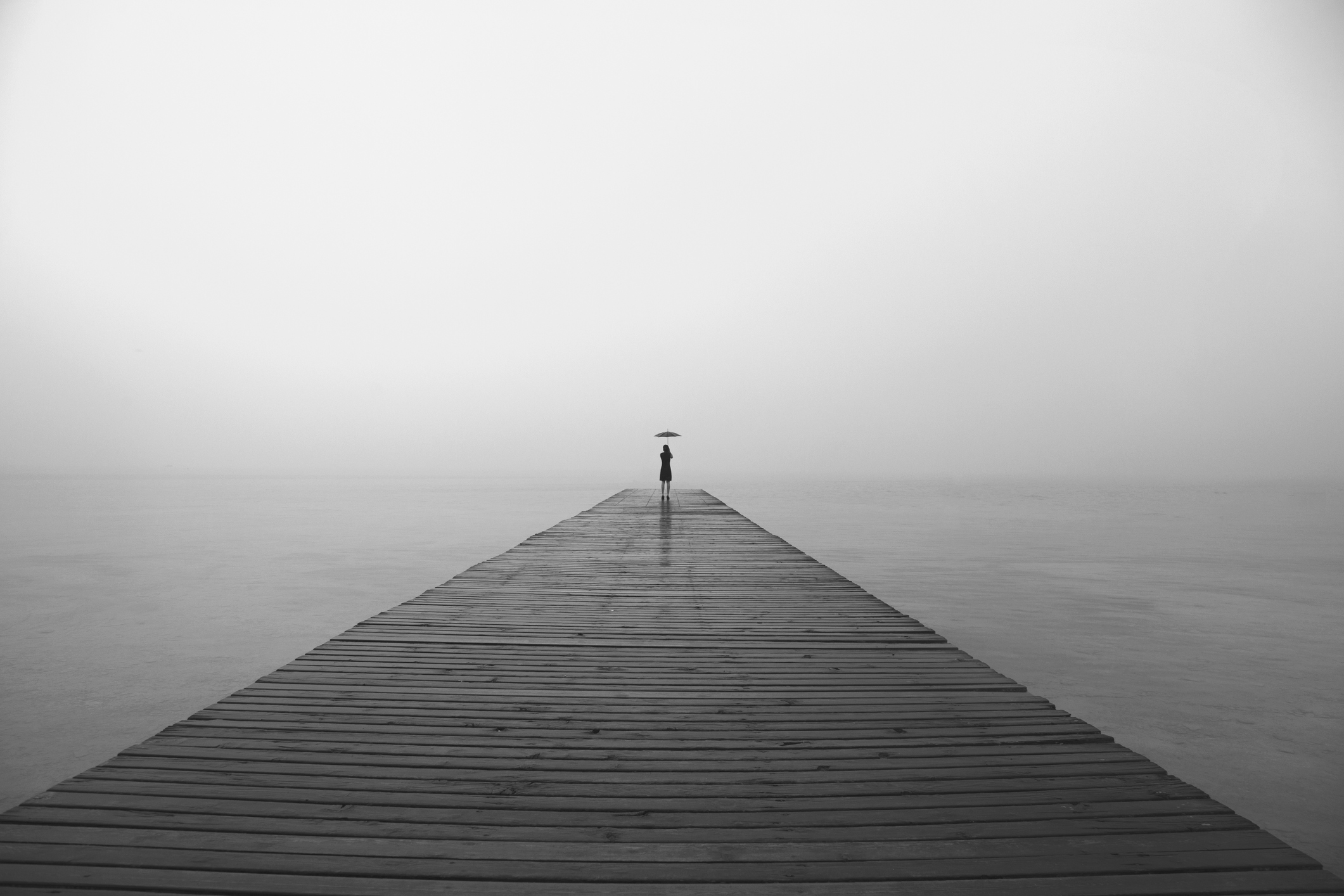 a long peer over water with a very small person at the very end with a black umbrella