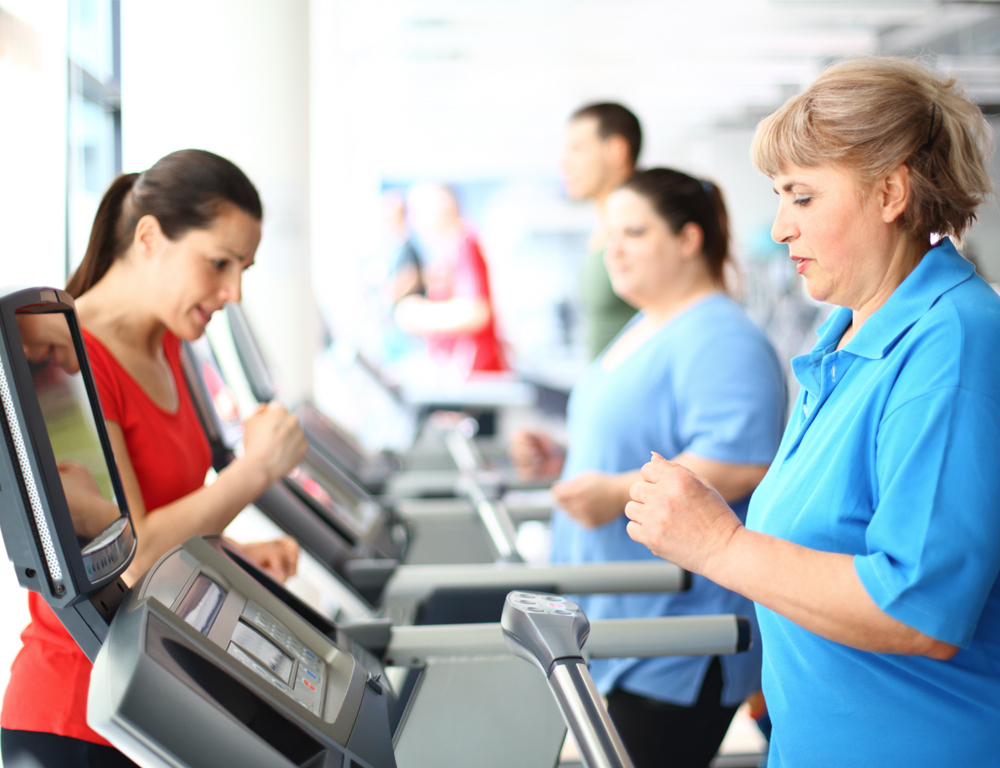 A woman in a blue shirt walks on a treadmill in the foreground. Other people are on treadmills in the background.