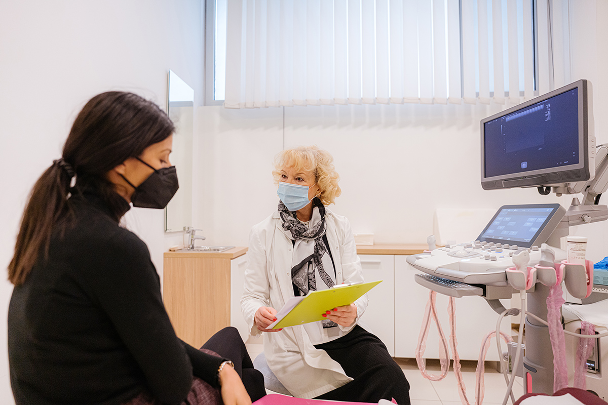 A patient wearing a mask has a conversation with a doctor wearing a mask in an exam room.