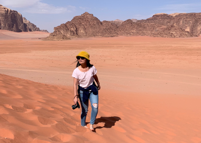 Young woman wearing sunglasses, a yellow hat, white t-shirt and jeans walks through the desert with a camera and stares off into the distance.