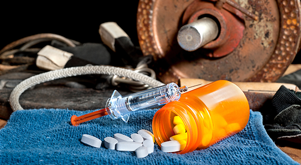 A pill bottle and syringe in the foreground; weights and other sports equipment are in the background.