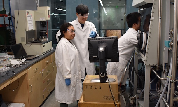 Two men and a woman in lab coats looking at polymer samples via a glove box and monitor.