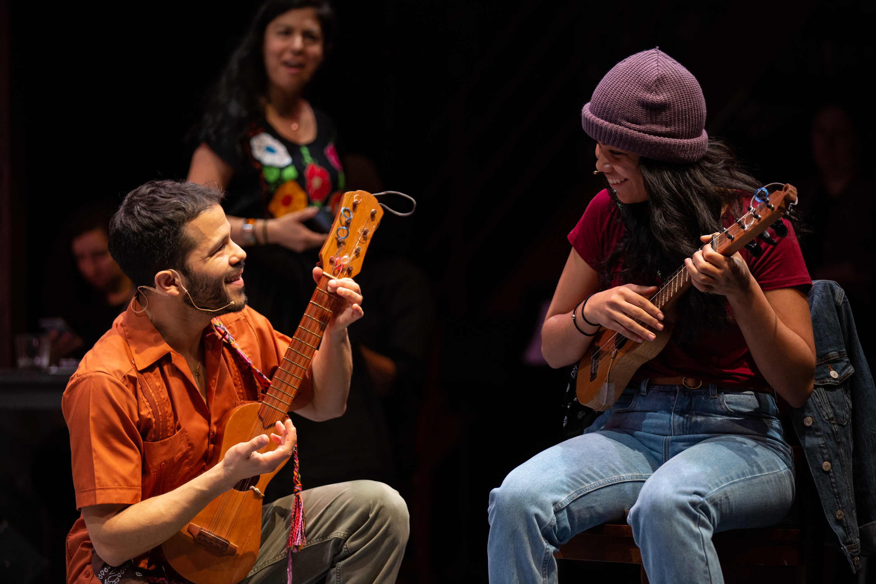 A man kneels next to a woman sitting in a chair while they both play stringed instruments.