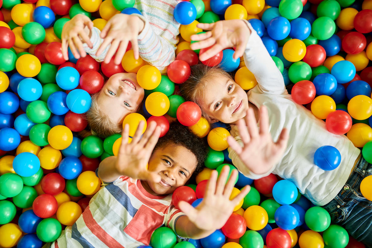 An overhead view of three young children laying in a pit of colorful balls with their hands extended into the air.