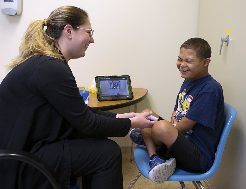 Whitney Adams, left, an intensive feeding therapist with the Penn State Children’s Hospital Feeding Program, holds out a toy to 11-year-old Jesus Rodriguez. They are both sitting in chairs at a small table. Adams is smiling. 