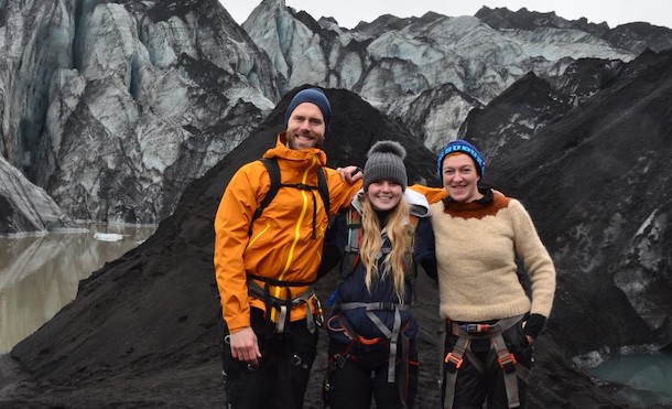 Three people standing on dark, rocky ground with rock formation behind them.