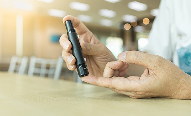 A close-up of a person's two hands shows the right hand holding a black device and using it to prick the pointer finger of his or her left hand. 