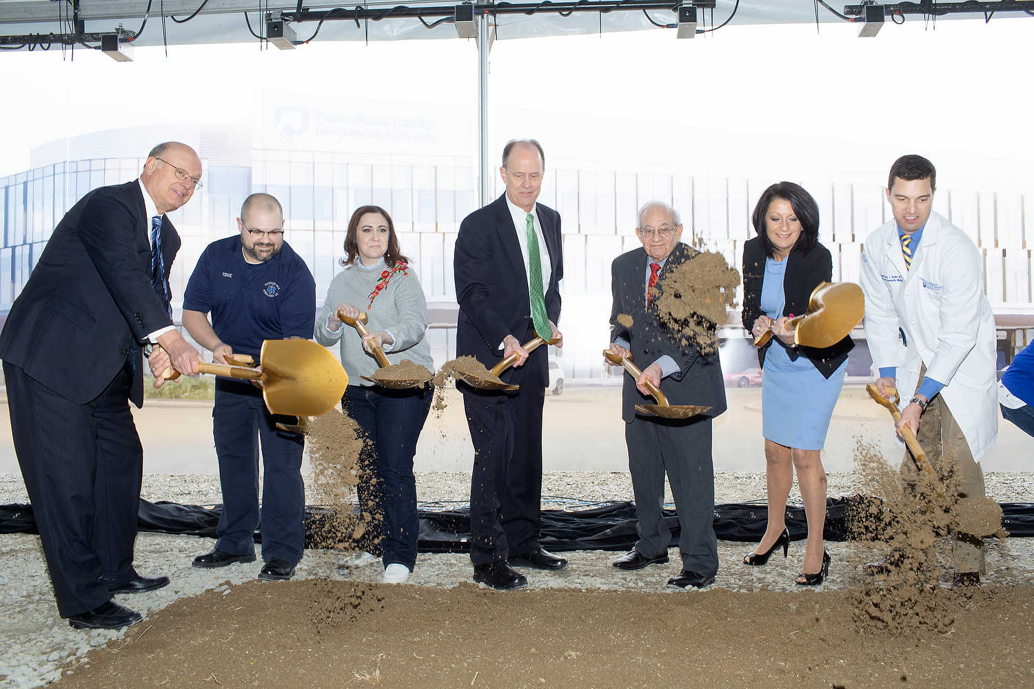 Seven people in formal attire hold gold shovels and toss dirt in the air. A large rendering of Penn State Health Hampden Medical Center is in the near background.