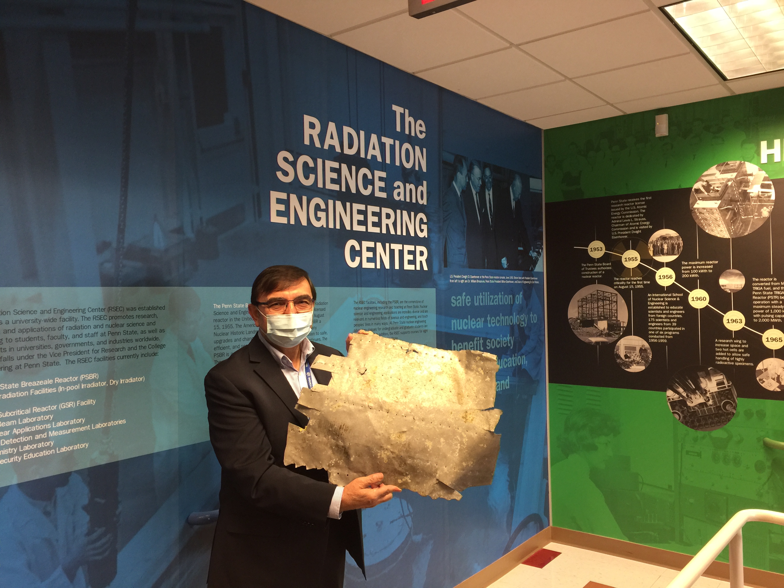 A man in a face mask and glasses stands in front of a blue wall that reads, "Radiation Science and Engineering Center," holding a piece of metal with rough edges and rivet holes. 