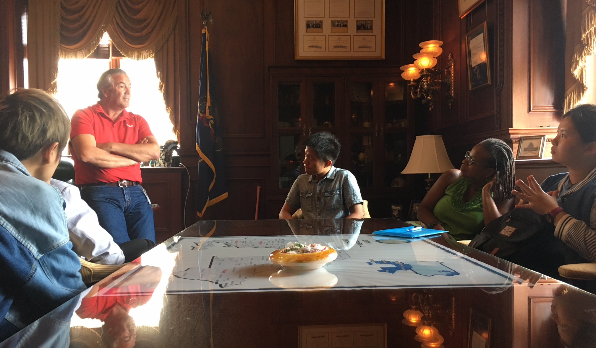 Five people talk around a large wooden conference table. Four are seated; a man who’s speaking is standing.