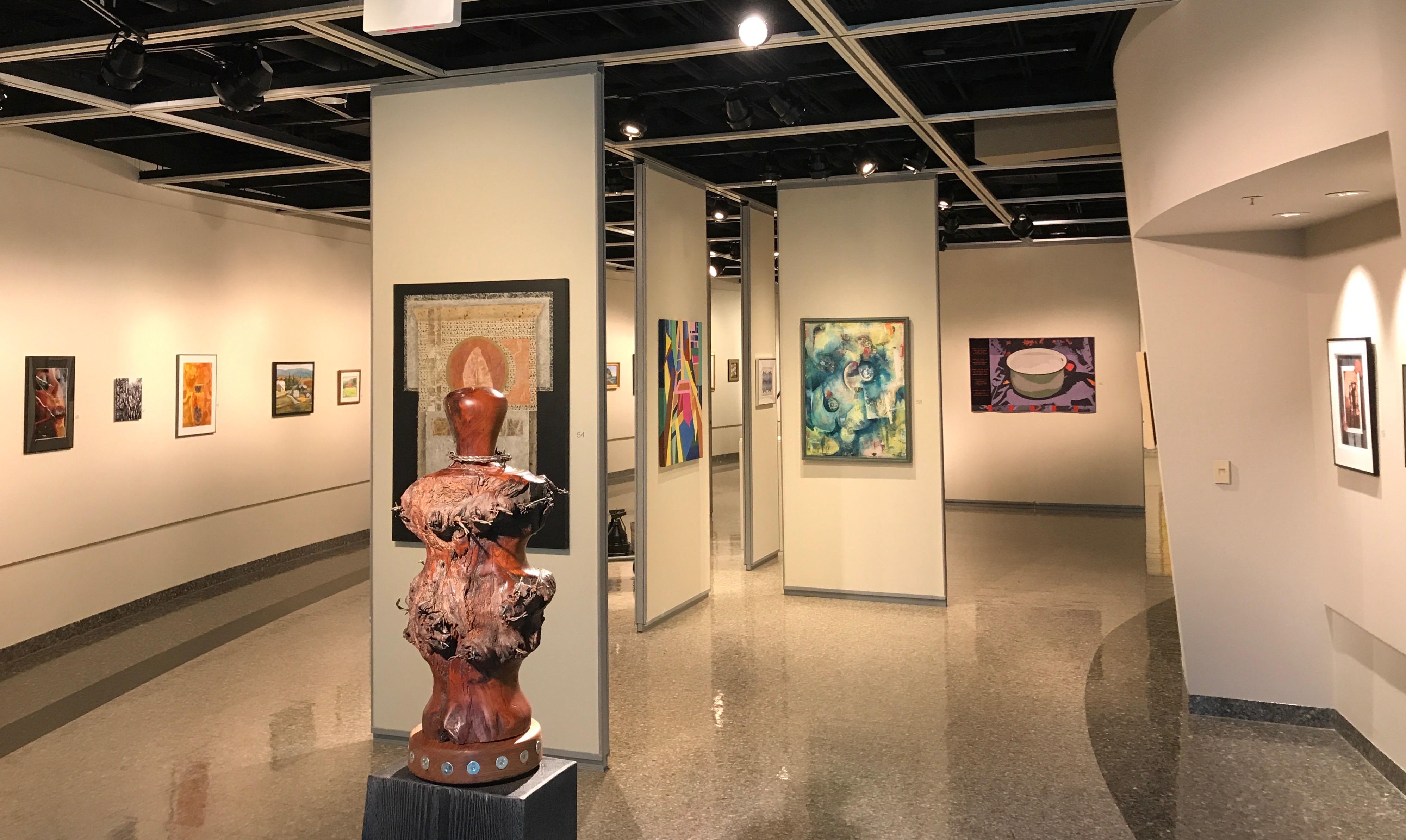 An overall photo of the Art Alliance exhibition in Robeson Gallery, showing a variety of paintings hanging on the walls with a large wooden sculpture in the foreground. 