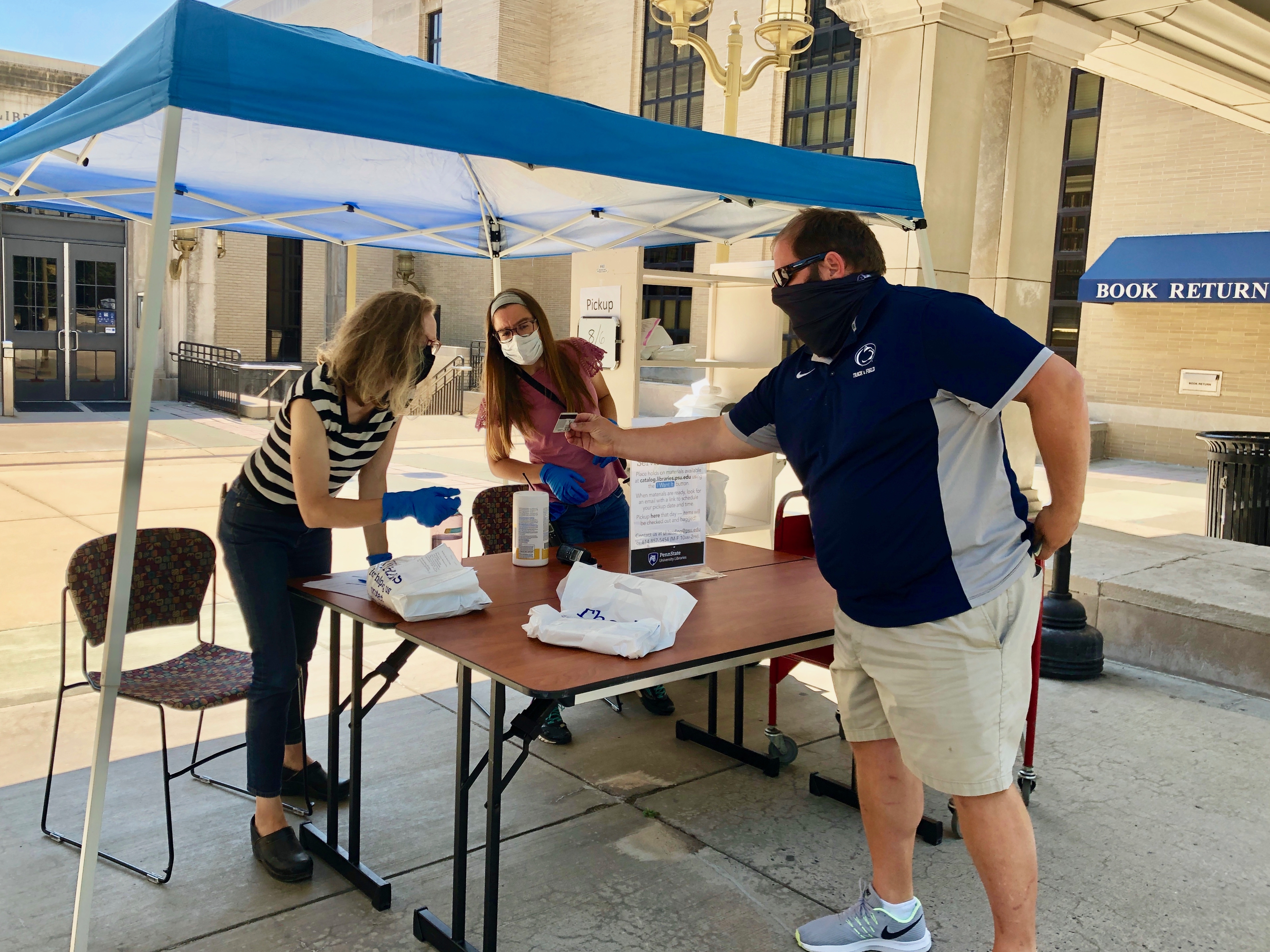 three people wearing masks under outdoor shade tent with man displaying ID card in hand to one woman wearing gloves while second woman observes