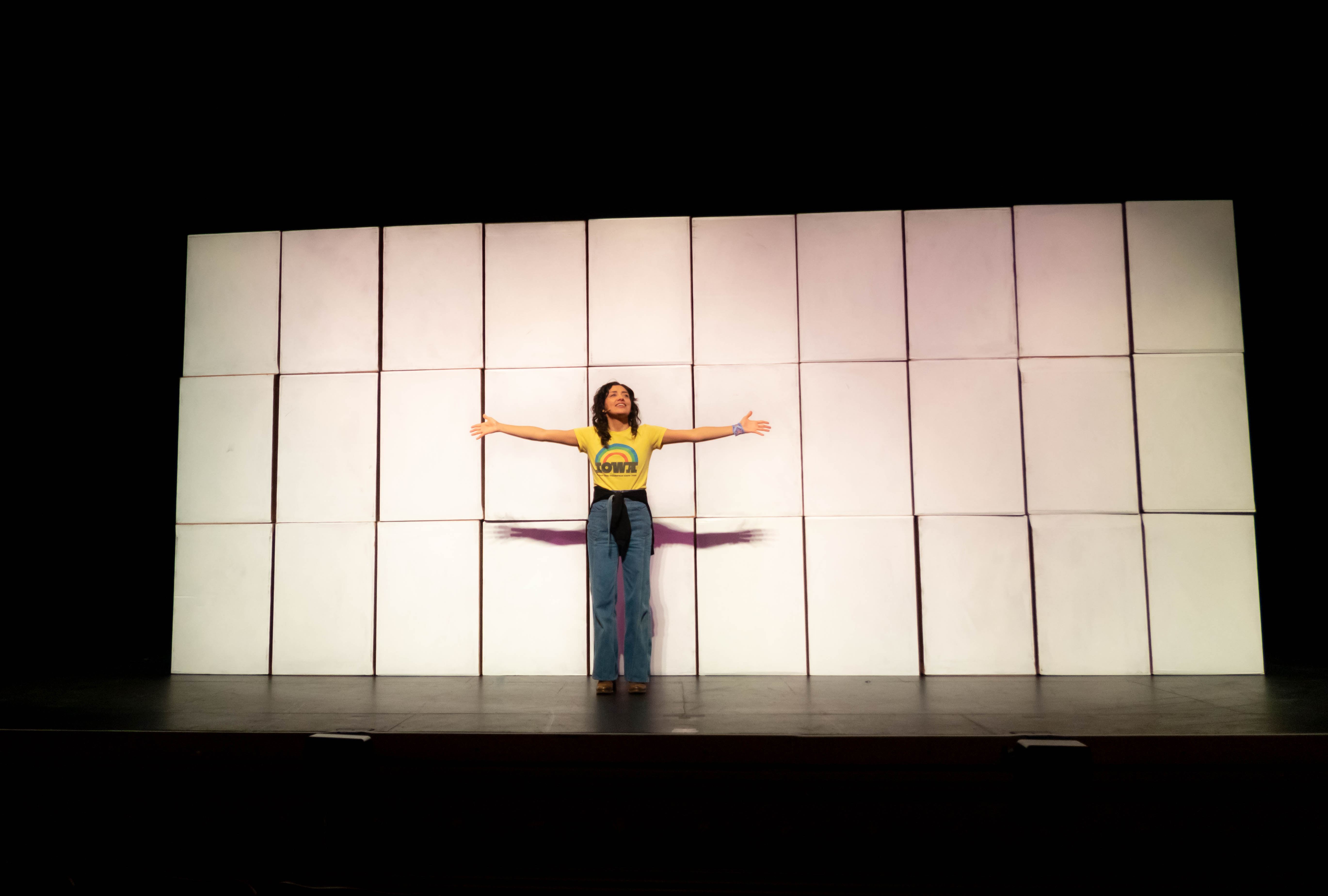 A woman stands in front of a wall of blank screens and stretches out her arms.