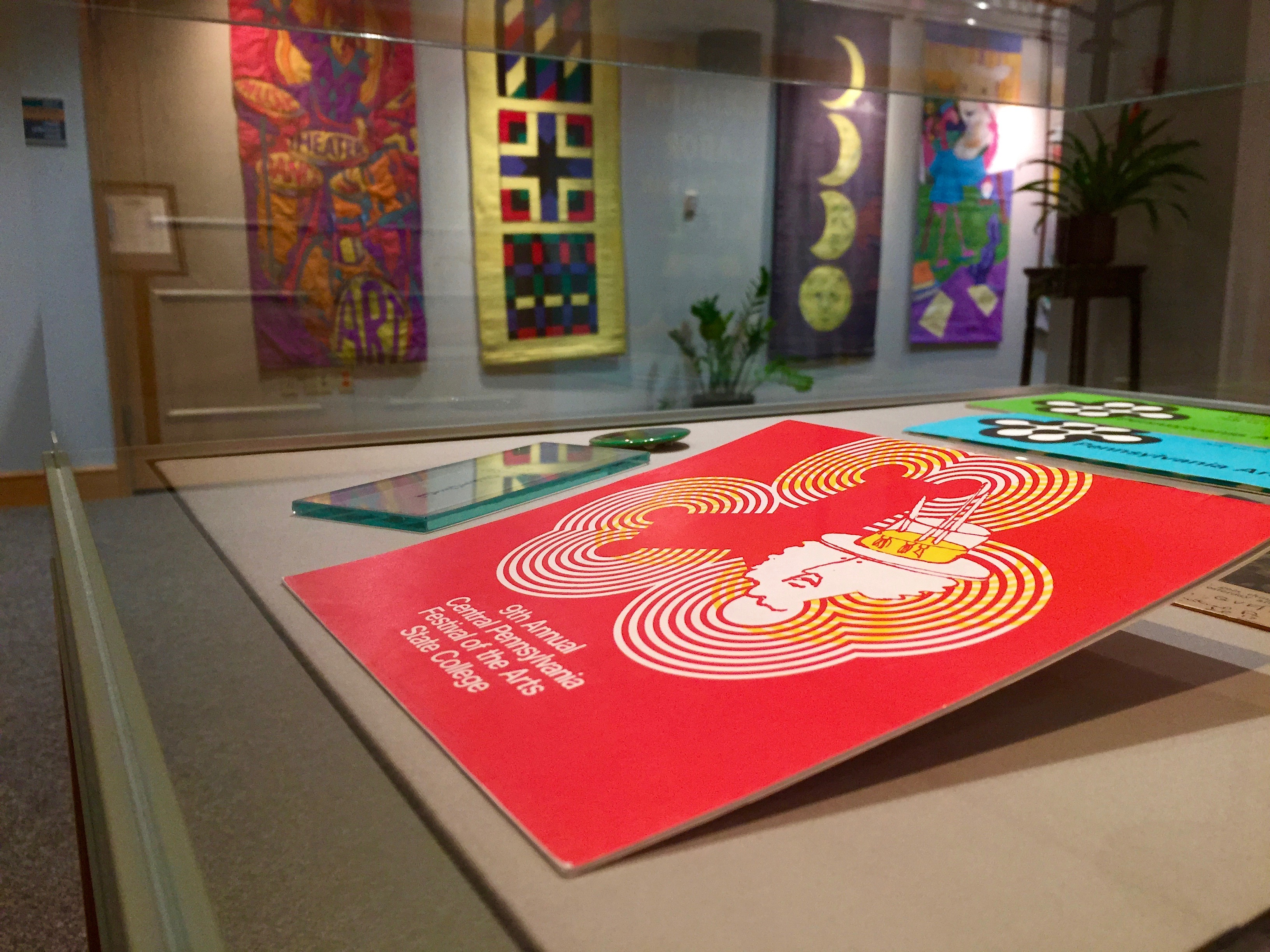 horizontal photo of glass exhibit case interior showing Arts Festival 9th annual red program covers and other artifacts with colorful banners hanging in background on wall