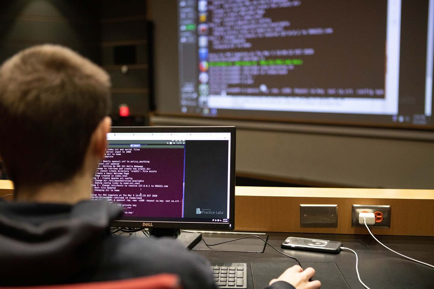 A male student codes on a computer screen while a teacher instructs on a large screen at the front of the classroom.