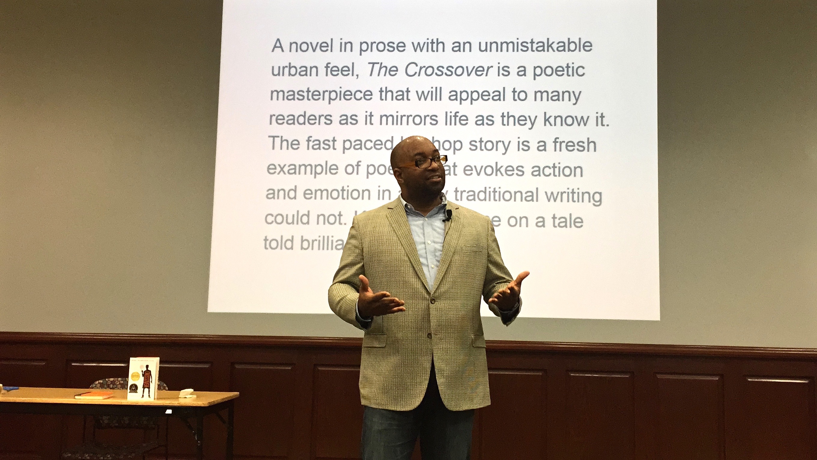 horizontal photo of Kwame Alexandar speaking to auditorium audience with review of his book "The Crossover" projected on wall behind him