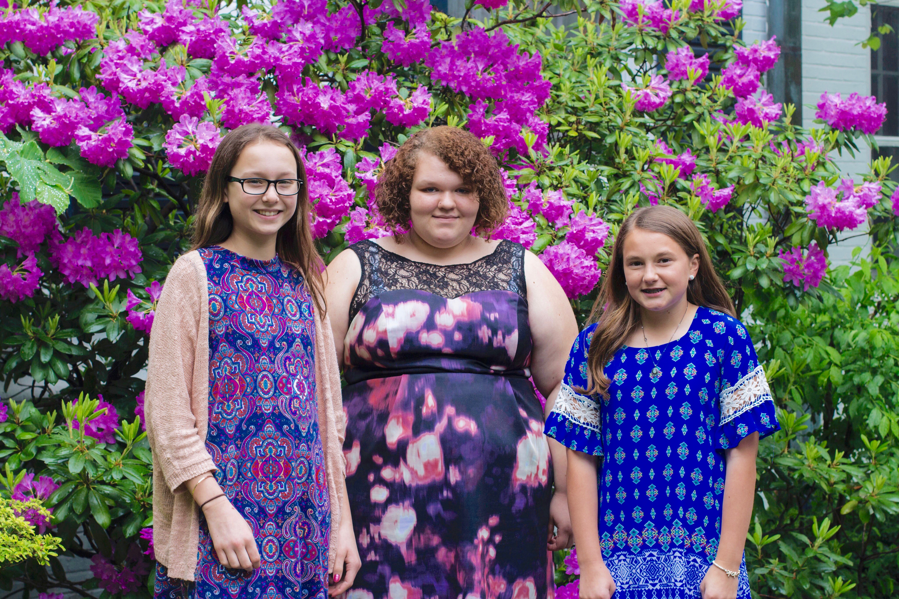 horizontal outdoor photo of three white female students standing in front of large blooming rhododendron shrub 