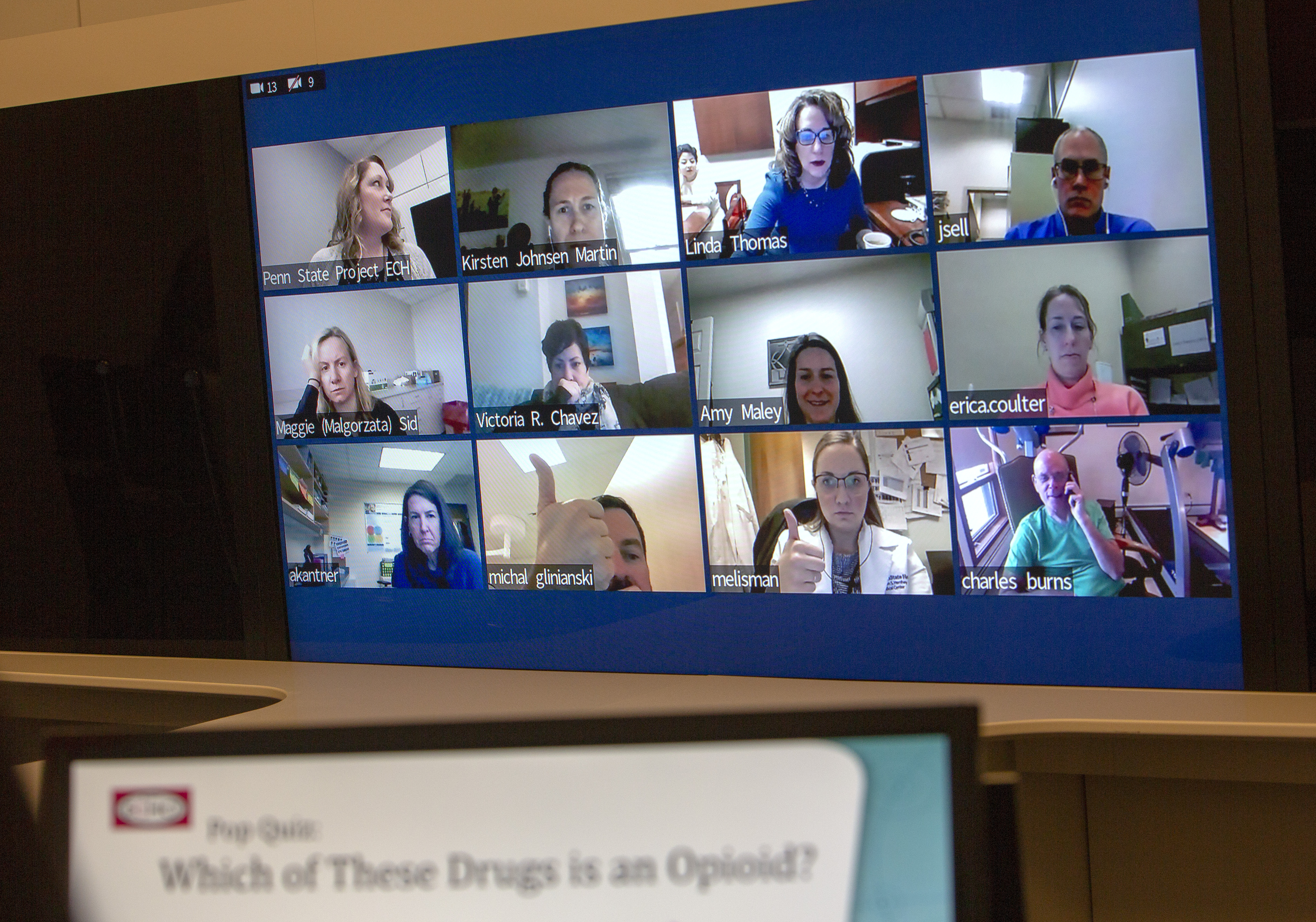 A large screen contains 12 mini screens showing physicians participating in Project ECHO. The participants are looking into their own web cams as they participate in the session. An out-of-focus screen reads “Pop Quiz: Which of these drugs is an opioid?”