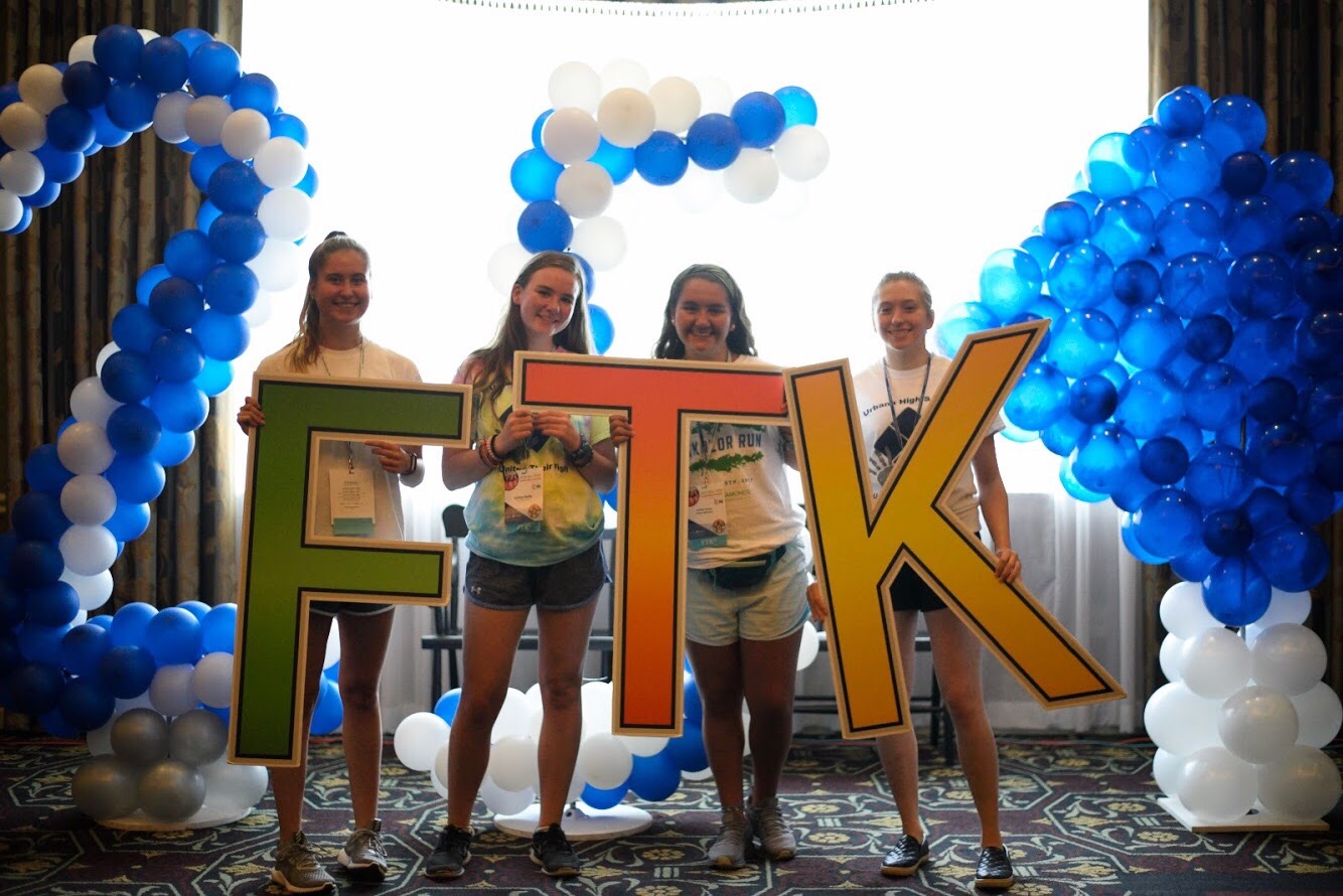 Four teenage girls hold oversized letters that spell out "FTK." Blue and white balloons and a window are in the background.