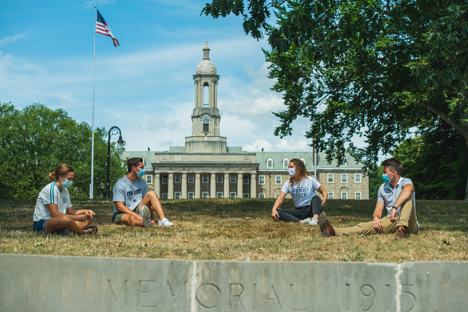 Four students sitting on the ground wearing masks with Old Main in the background on a clear day