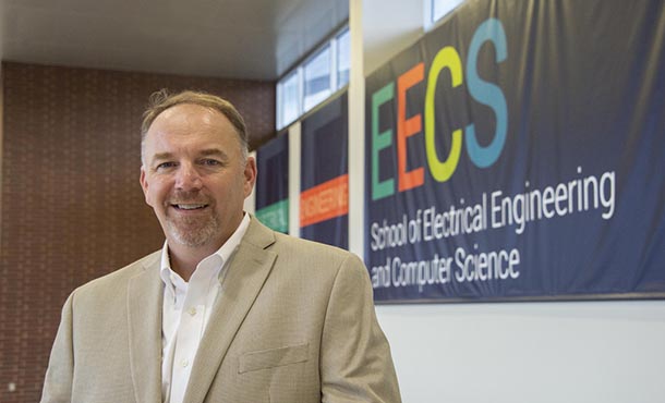 A man in a tan suit jacket poses in front of a School of EECS wall banner