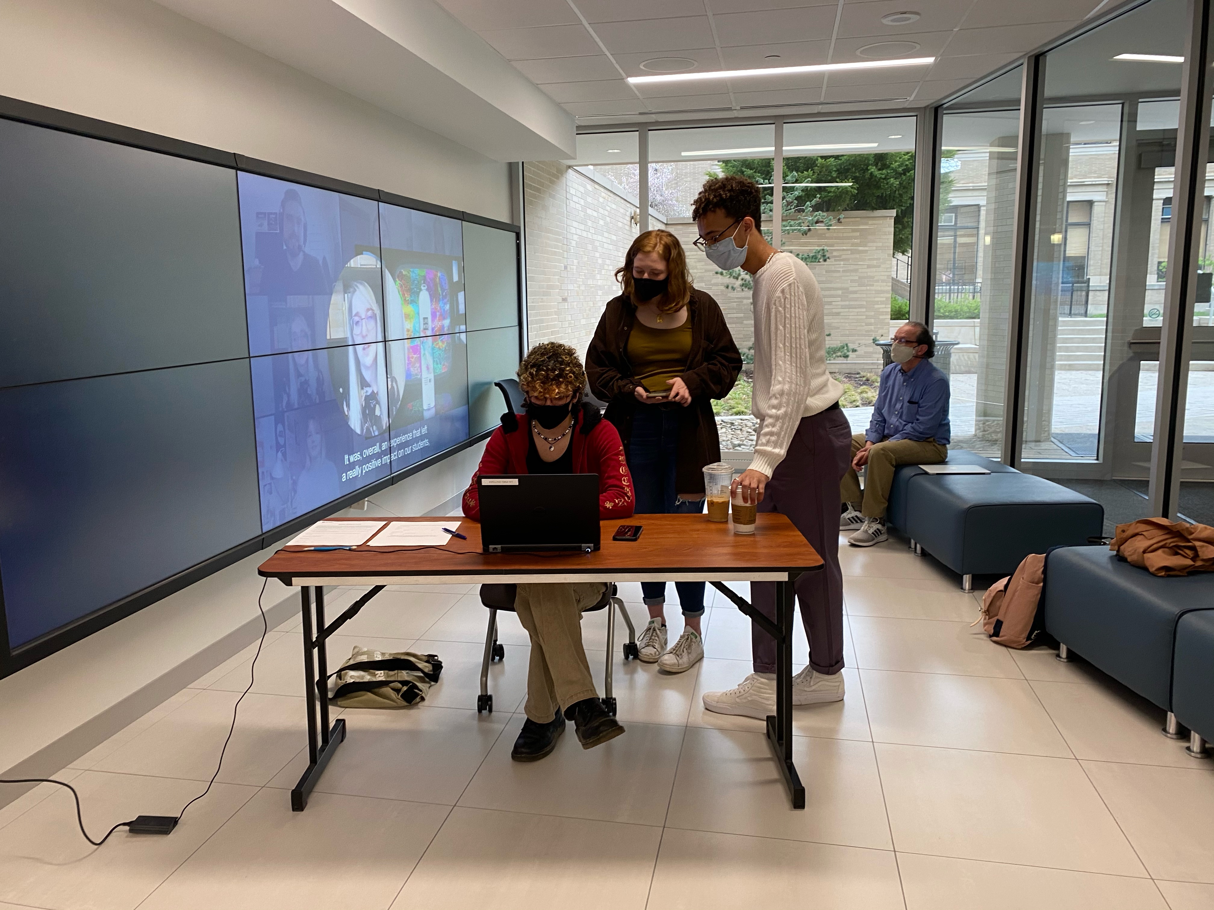 two masked students standing behind seated masked student in front of laptop and masked male seated in background